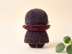 A small crocheted plush doll shown from the back, wearing a dark charcoal-gray outfit and a burgundy collar, set against a warm cream background with a green leaf prop in the corner.