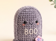 A cute, hand-crocheted plush ghost toy in a textured lavender-gray yarn. It features embroidered black eyes and a smiling mouth, pink cheeks, the word 'BOO' stitched in cream on its belly, and a small pink and purple flower on the bottom right.