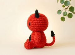 A crocheted devil doll in bright red with black horns, tail tip, and belly patch, sitting on a warm cream background with a green leafy branch in the upper right corner.