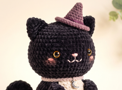 A close-up of a crocheted black cat toy wearing a purple witch hat and a cream-colored collar with a small silver crescent moon charm, set against a soft beige background.