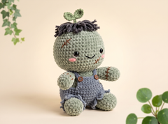 A cute crocheted plush toy with a round light green head, dark gray yarn hair with two green leaves on top, pink stitched cheeks, and blue-gray overalls, posed against a soft beige background with decorative green leaves in the corners.