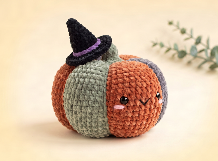 A cute crocheted pumpkin plush toy with a black witch hat and purple band, featuring orange, sage green, and gray sections, set against a warm beige background with a soft-focus eucalyptus branch.