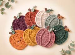 A set of crocheted pumpkin-shaped items in multiple autumn colors, including orange, yellow, pink, green, purple, and terracotta, arranged on a light beige background with decorative eucalyptus leaves.