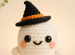 A close-up of a crocheted white ghost doll wearing a black witch hat with an orange band, set against a soft beige background.