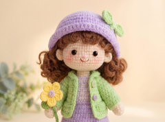A crocheted doll with curly brown hair, wearing a lavender hat with a green bow, a green cardigan, and holding a yellow flower, set against a soft beige background.