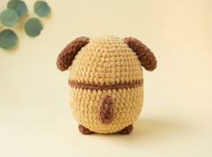 A crocheted plush toy shaped like a small dog or bunny, shown from the back. The body is made of textured beige yarn with dark brown accents for the ears, tail, and legs, set against a soft beige background with a decorative eucalyptus leaf in the upper left corner.