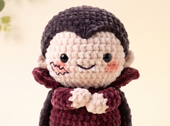 A close-up of a crocheted doll with a pale face, dark brown hair, pink cheeks, and a burgundy dress against a soft cream background.