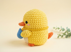 A yellow crocheted duck toy with orange beak and feet, holding a blue egg, set against a warm beige background with a small green eucalyptus sprig on the right.