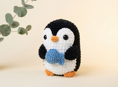 A crocheted penguin toy holding a blue fish, standing on a light beige surface with a soft beige background and a decorative eucalyptus branch in the upper left corner.