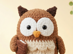 A close-up of a crocheted owl toy or plush figure with a brown body, large white eyes, a small orange beak, and dark brown ears, holding a brown mug against a soft beige background.