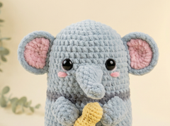 A crocheted elephant toy in light blue-gray yarn with pink inner ears, black bead eyes, and a yellow banana held in its paws, set against a soft beige background.
