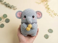 A crocheted plush elephant toy held in a person's hand against a warm cream background with scattered eucalyptus leaves. The elephant is light blue-gray with pink inner ears, black eyes, and a small yellow peanut held in its front hooves.