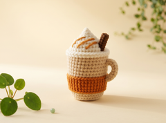 A crocheted mug-shaped dessert or drink cozy with white whipped cream, orange drizzle, and a brown cookie decoration, set on a warm beige background with soft green plant leaves.
