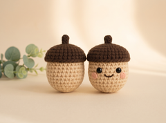 Two crocheted acorn toys with smiling faces, one plain and one with embroidered cheeks, set against a warm beige background with a small green plant in the side.