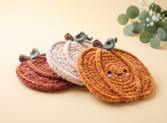 Three crocheted pumpkin-shaped coasters with cute embroidered faces, displayed on a warm beige background with a decorative eucalyptus sprig in the corner.