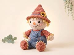 A crocheted scarecrow doll with a brown hat, blue overalls, and a beige face, posed against a soft cream background with small green leaves in the corners.
