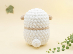 A small white crocheted plush sheep toy with tan ears and a tan band around its middle, photographed on a soft cream background with decorative green leaves in the corners.