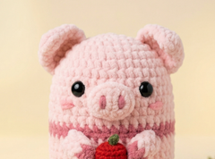 A pink crocheted plush pig toy holding a small red apple, photographed against a soft warm beige background.