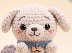 A close-up of a crocheted plush puppy toy with floppy ears, large black eyes, pink stitched cheeks, a blue-gray collar, and a small wooden bone-shaped tag, set against a soft beige background.