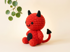 A crocheted devil doll in bright red with black horns, tail tip, and belly patch, sitting on a warm cream background with a green leafy branch in the upper left corner.
