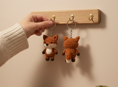 A hand wearing a cream knit sweater holds one side of a crocheted fox keychain next to a wooden wall hook display. The keychain features a brown and orange fox with a white chest and dark brown paws and tail tip, shown from both front and back views.