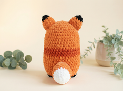 A crocheted fox toy shown from the back, featuring an orange and brown striped body, a white tail, and black ear tips, set against a warm beige background with soft green eucalyptus leaves.