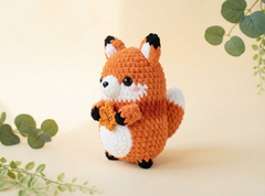 A crocheted fox toy holding an orange leaf, standing on a light beige background with decorative green leaves in the corners.