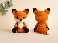 A crocheted fox toy shown from the front and back, made with orange, white, and black yarn, set against a warm beige background with a small eucalyptus sprig in the corner.