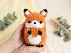 A hand holding a crocheted fox plush toy with orange, white, and black details, set against a soft beige background with decorative eucalyptus leaves.