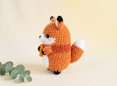 A crocheted fox toy in orange, white, and black, standing on a light beige background with a sprig of green eucalyptus leaves in the lower left corner.