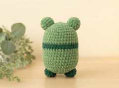 A small crocheted plush toy shaped like a rounded character with two pom-pom ears, shown from the back on a light wooden surface with soft-focus greenery and a cream background.