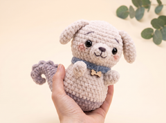 A hand holding a crocheted plush puppy toy with a cream-colored body, floppy ears, a blue collar, and a small wooden bone charm, set against a soft beige background with blurred green leaves in the corner.