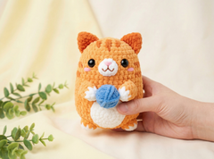 A small crocheted orange cat toy holding a blue ball of yarn, being held by a human hand against a soft cream fabric backdrop with a green plant in the corner.