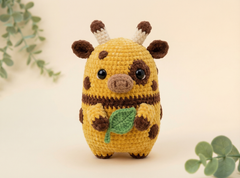 A crocheted plush toy shaped like a giraffe pig, featuring a yellow body with brown spots, a brown snout and ears, white and brown horns, and a green leaf in its front hooves, set against a warm beige background with soft green foliage accents.