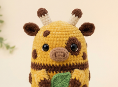 A close-up of a crocheted giraffe plush toy with a yellow body, brown spots, cream horns, and a green leaf detail, set against a soft beige background.