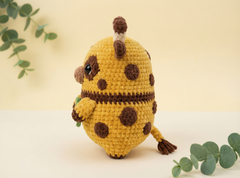 A crocheted giraffe toy in mustard yellow and brown, standing upright on a light cream surface against a pale yellow background with decorative eucalyptus leaves.