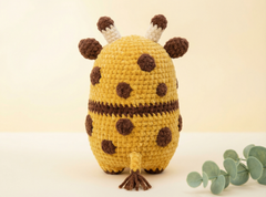 A crocheted plush giraffe toy shown from the back, featuring a yellow body with dark brown spots and stripes, cream and brown ossicones, and a small tail, photographed on a light beige background with a sprig of eucalyptus at the lower right.