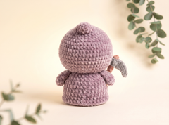 A handmade crocheted doll in a dusty lavender-purple color, shown from the back holding a small gray scythe, set against a warm cream background with soft eucalyptus leaves.