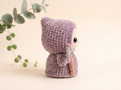 A small crocheted doll with a lavender-purple hood and matching body, holding a gray staff, photographed on a warm cream background with a green trailing plant at the side.