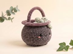 A cute crocheted pot-shaped basket in dark gray with a dusty purple handle and rim, decorated with small green crochet flowers and embroidered facial features, set against a warm beige background with decorative green leaves.