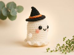 A cute crocheted ghost doll wearing a black witch hat with an orange band, set against a warm beige background with soft green plant leaves in the corners.