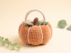 A crocheted pumpkin-shaped handbag with a cream handle, cream vertical stripes, a brown stem, and green leaves, displayed on a warm beige background with decorative eucalyptus leaves.