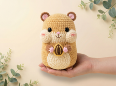 A crocheted hamster toy held in a person's hand against a warm cream background with soft green eucalyptus leaves.