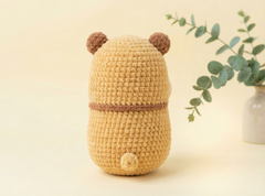 A crocheted plush toy shaped like a small bear or pig, shown from the back. The toy is made of textured tan yarn with brown ears and a horizontal band around the middle, set against a warm cream background with a blurred green plant in a vase on the right.