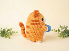 A crocheted orange tabby cat plush toy holding a blue ball, photographed on a warm cream background with small green eucalyptus sprigs on both sides.
