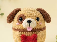 A close-up of a crocheted plush dog toy with tan and cream fur, brown floppy ears, a pink tongue, and a red bone-shaped accessory in front, set against a soft beige background.