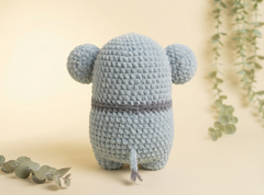 A light blue crocheted plush toy shaped like a small animal with round ears and a short tail, shown from the back on a warm cream background with eucalyptus leaves.