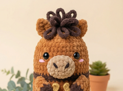 A crocheted plush toy resembling a cute brown animal character with a dark brown hair accessory, set against a warm beige background with a small potted succulent in the background.