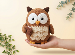 A crocheted owl plush toy with large white eyes, a yellow beak, and brown and cream feathers, being held in a person's hand against a warm beige background with decorative green leaves.