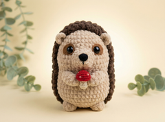 A crocheted hedgehog toy holding a red mushroom, centered against a warm beige background with soft green eucalyptus leaves in the corners.